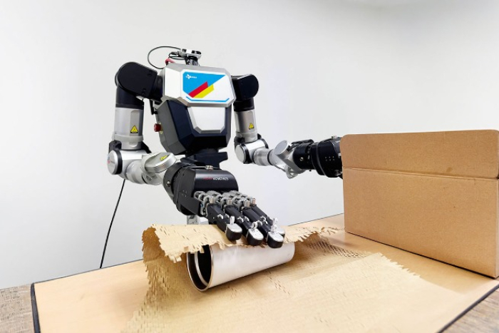Humanoid with a multi-finger hand wrapping a tumbler in kraft paper next to a cardboard box