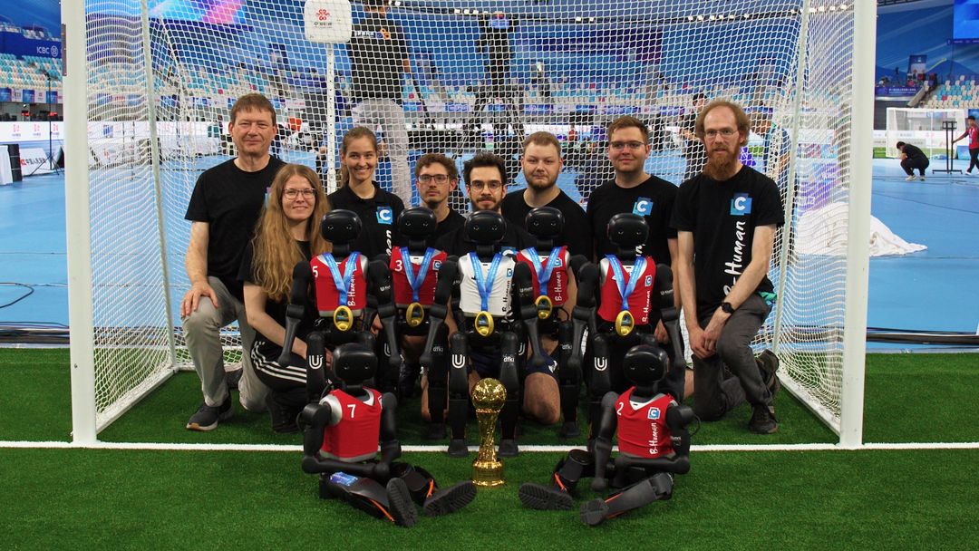 B-Human researchers and medal-wearing NAO-class robots posed in a goal with a trophy