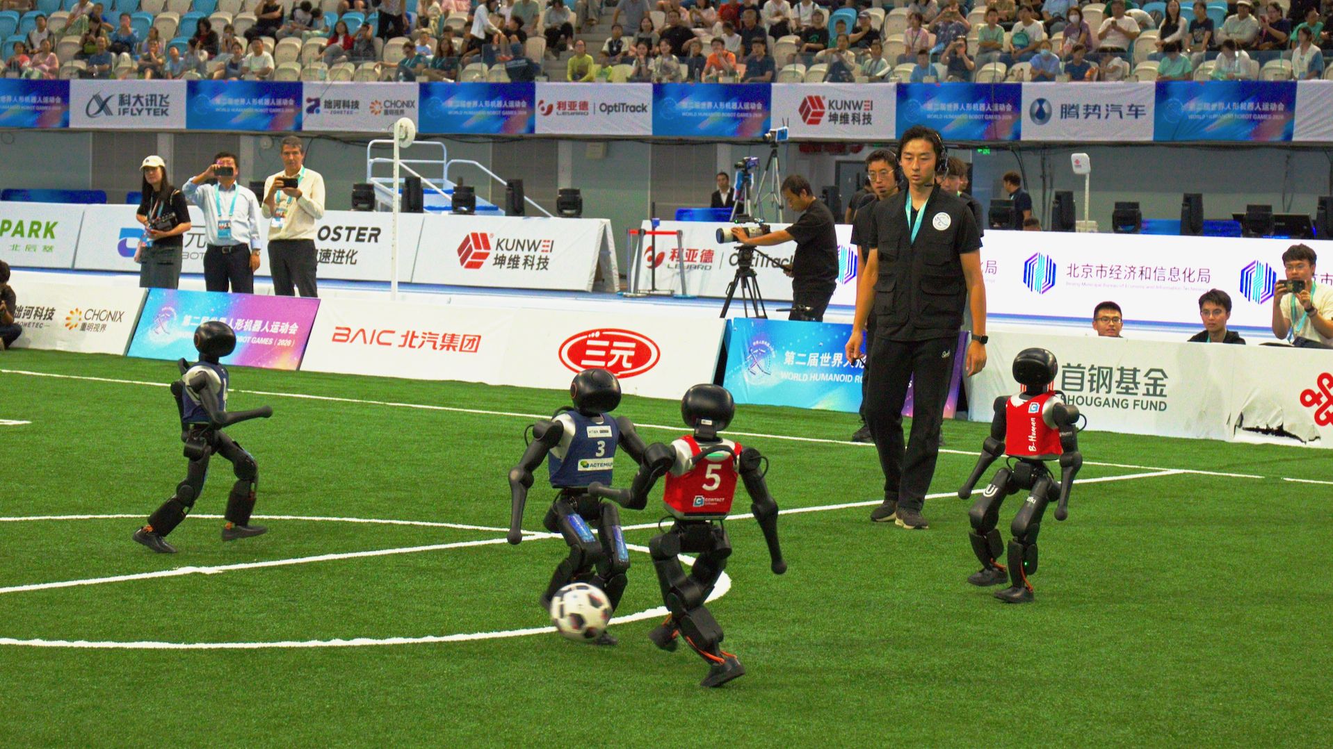 B-Human robot in a red number-5 jersey kicking a soccer ball against blue-jersey opponents on a turf pitch in Beijing