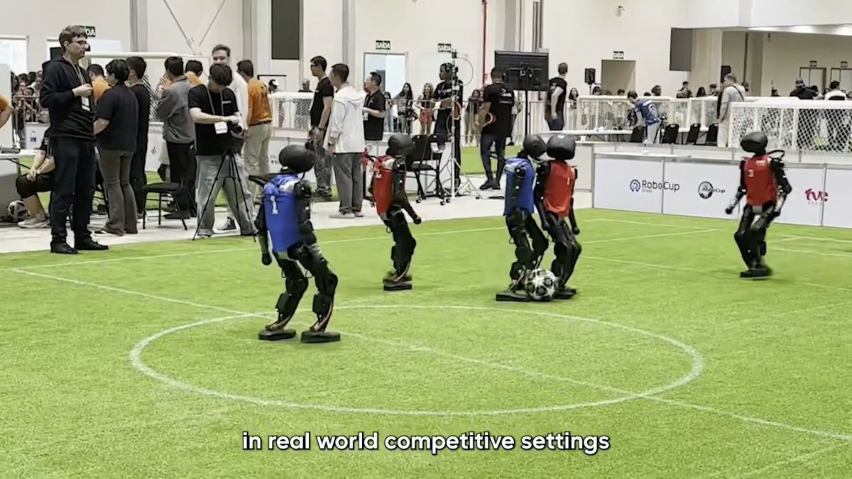 Booster T1 humanoids in RoboCup jerseys on an indoor soccer pitch