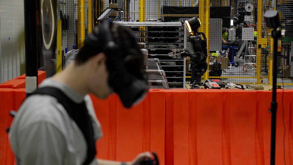A person wearing a headset stands near an Atlas robot in a caged work cell