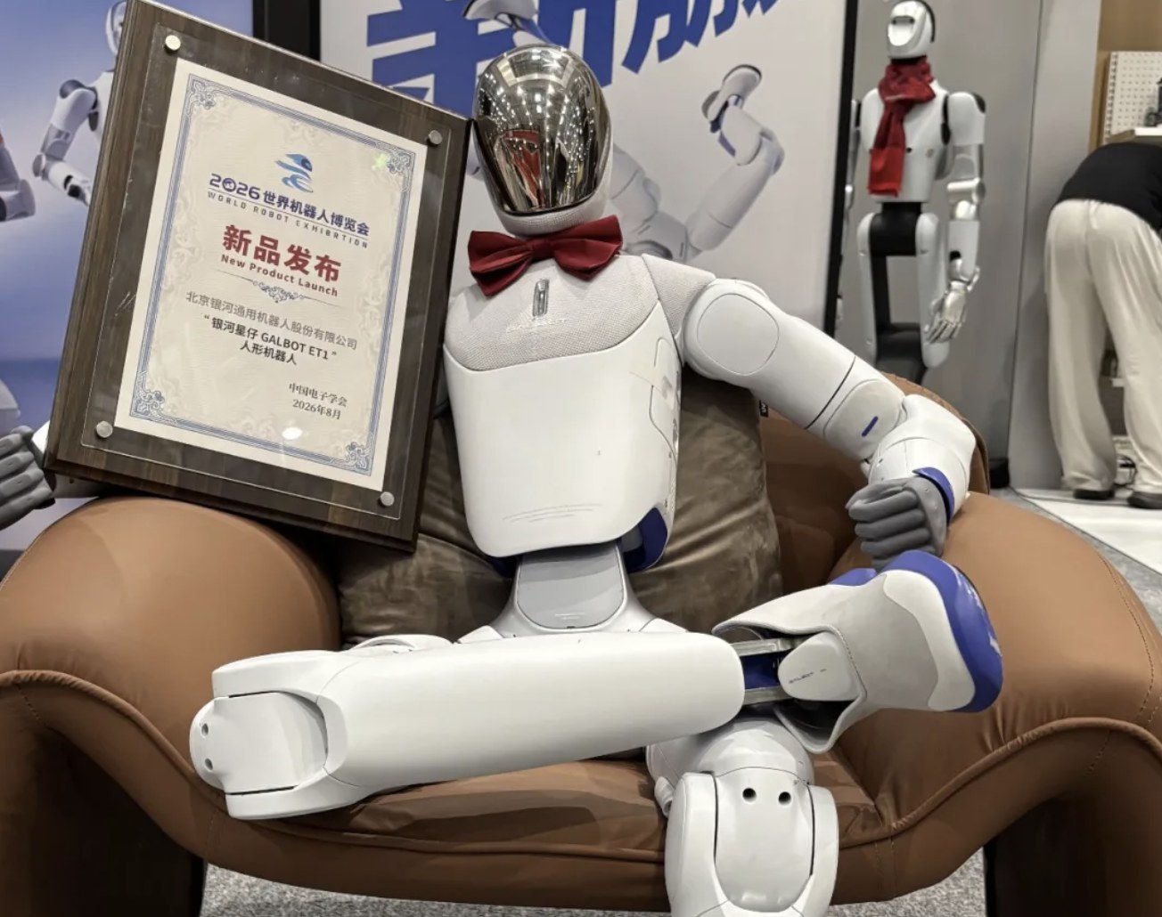 White Galbot ET1 sitting in an armchair holding a 2026 World Robot Exhibition new-product plaque, bow tie on