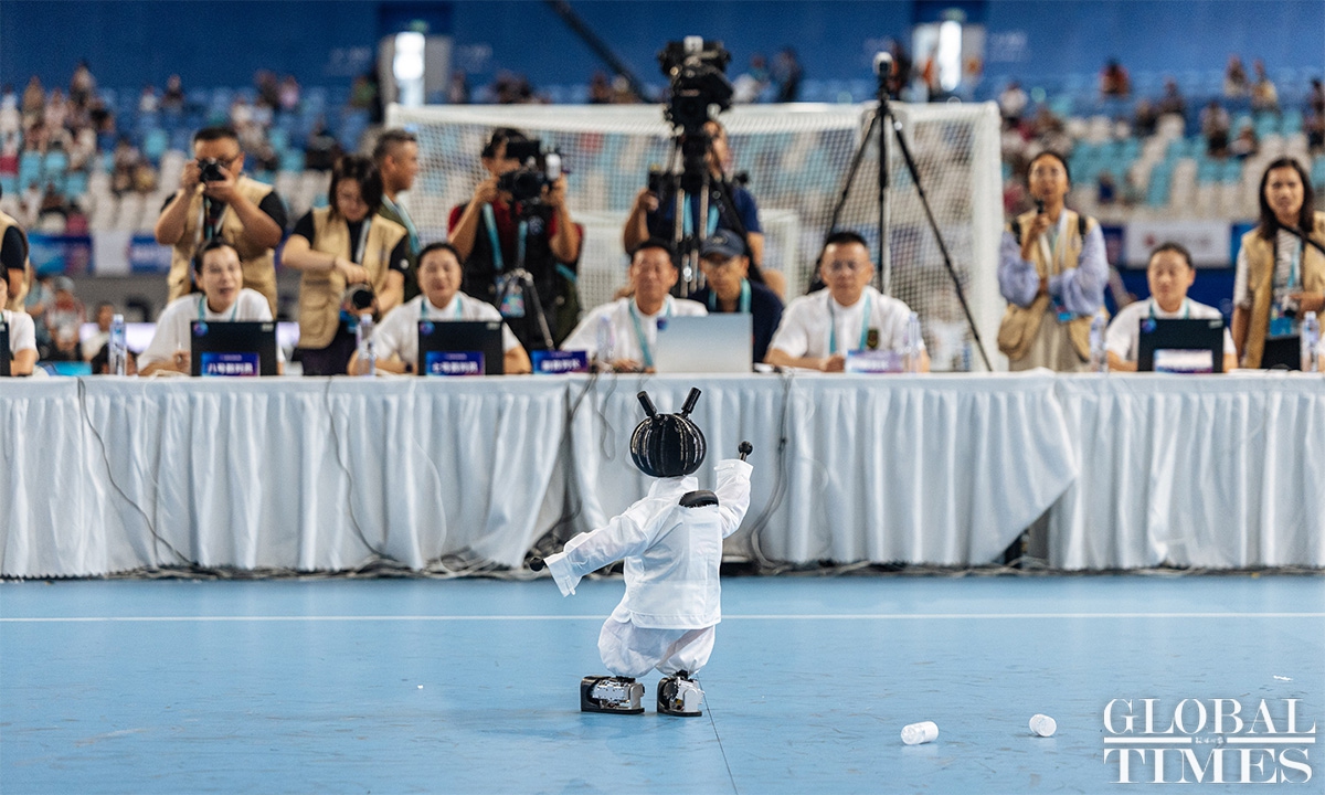 Small humanoid in a white jacket performing in front of a long judges’ table