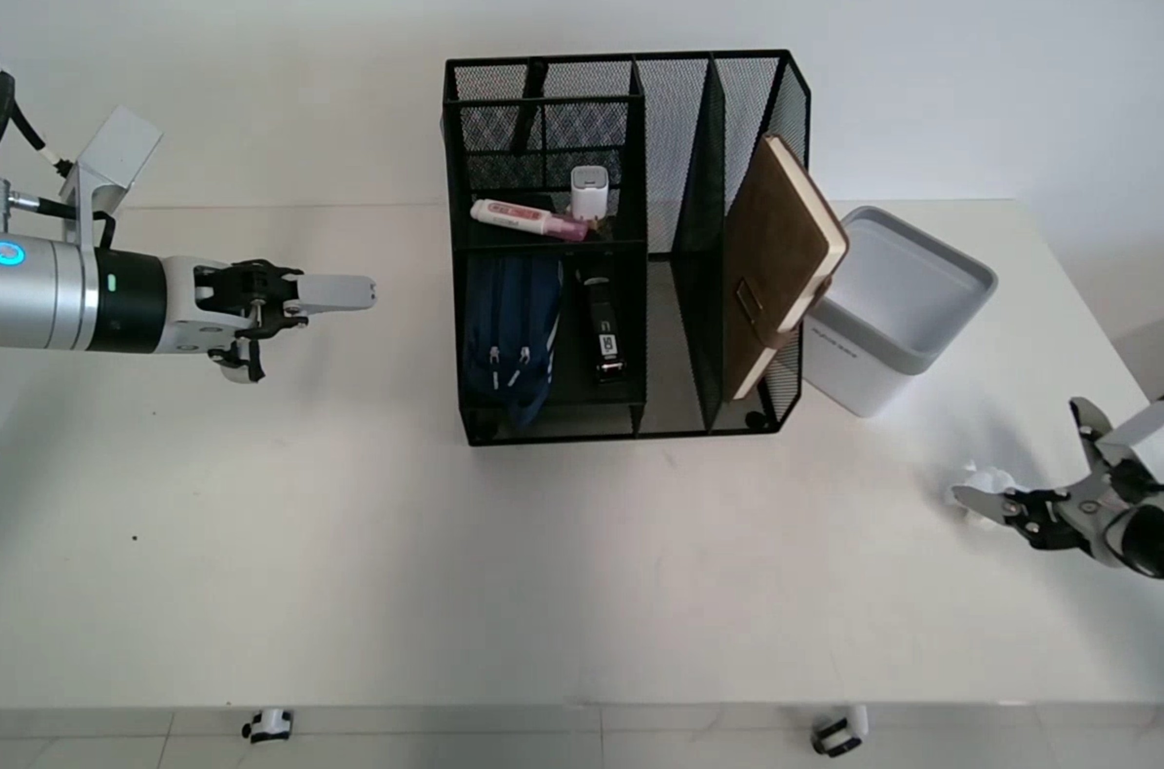 Unitree G1 arms on a white desk with a mesh organizer, tissue box, and objects for a pick-and-place task