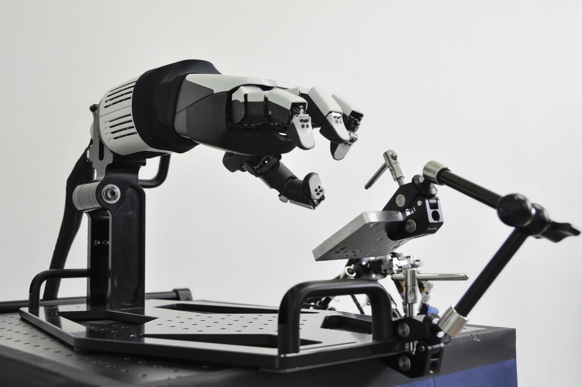 Honda four-finger robotic hand on a lab fixture reaching toward a metal workpiece