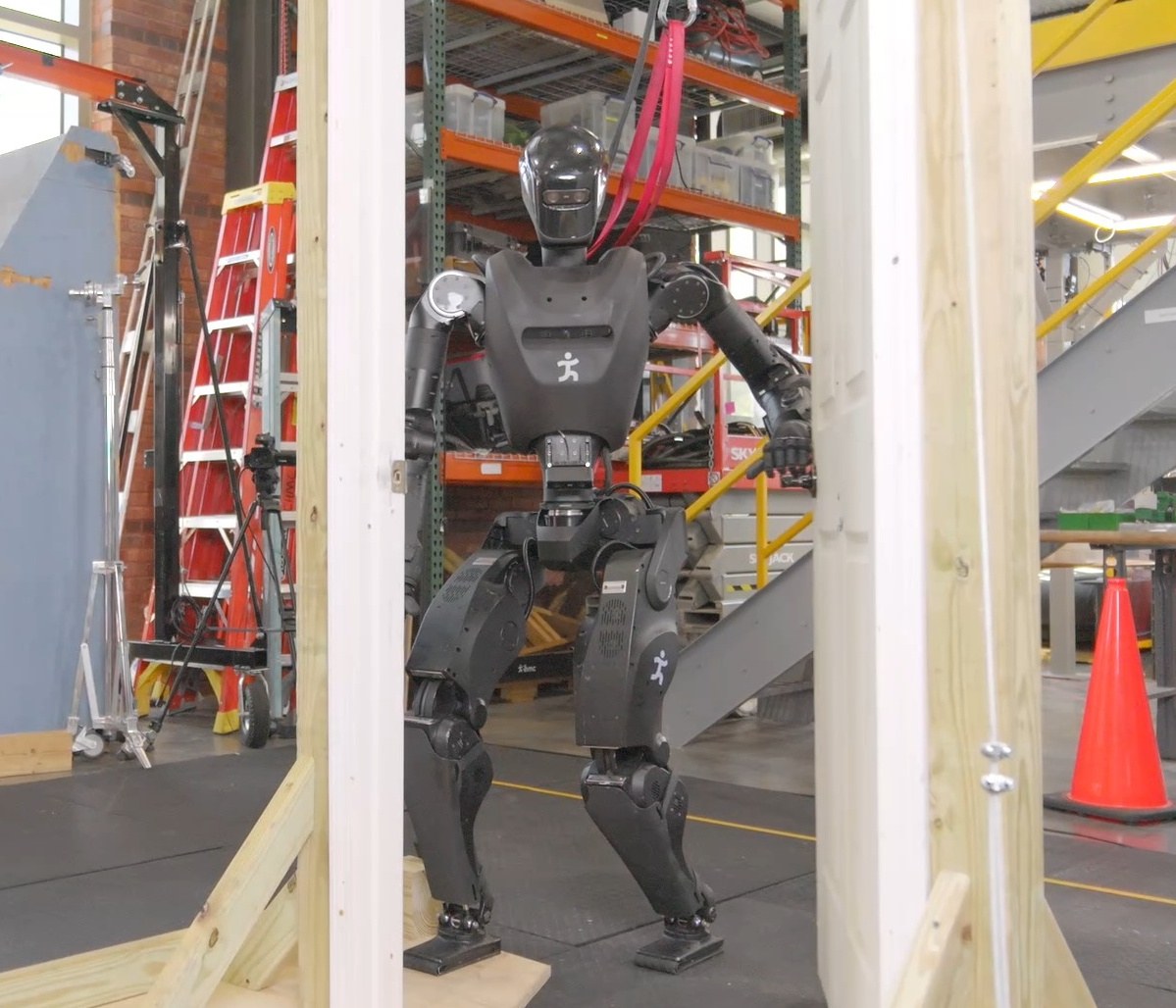 Black Alex humanoid walking through a wooden door frame in a lab, safety gantry overhead