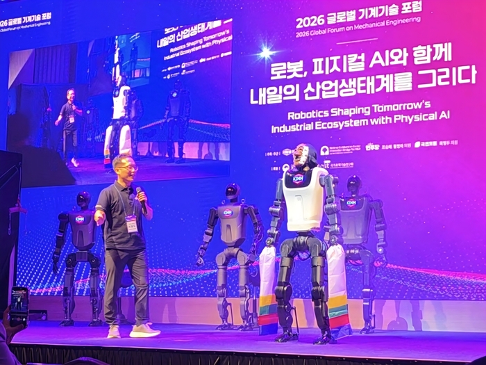 Presenter on stage with several KAIROS units, one wearing hansam, under a Physical AI forum backdrop