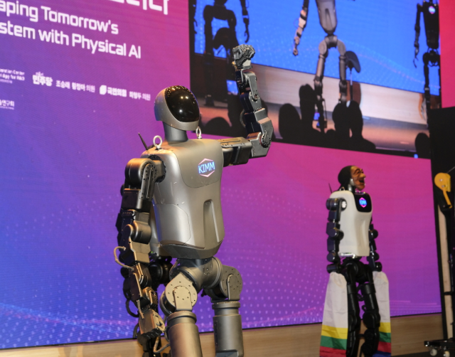 KAIROS humanoid with KIMM on its chest waving on stage, another unit in a mask and hansam sleeves behind