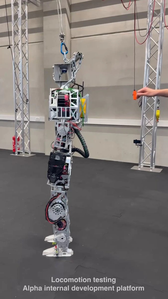 Boxy silver humanoid prototype hanging from a gantry in a warehouse, labeled locomotion testing alpha internal development platform
