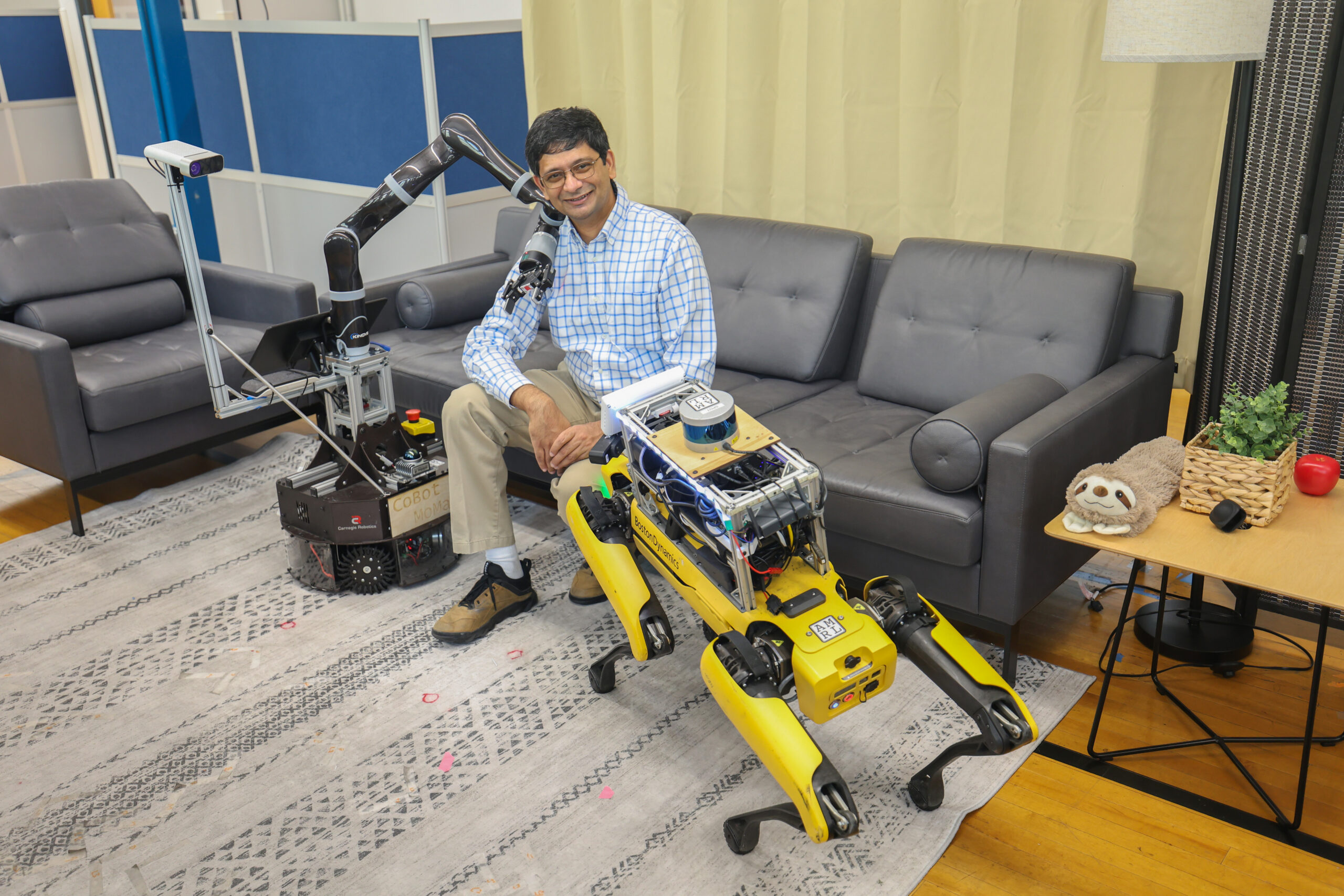 Joydeep Biswas seated on a sofa in a UT lab with a Boston Dynamics Spot quadruped and a CoBot MoMa mobile manipulator