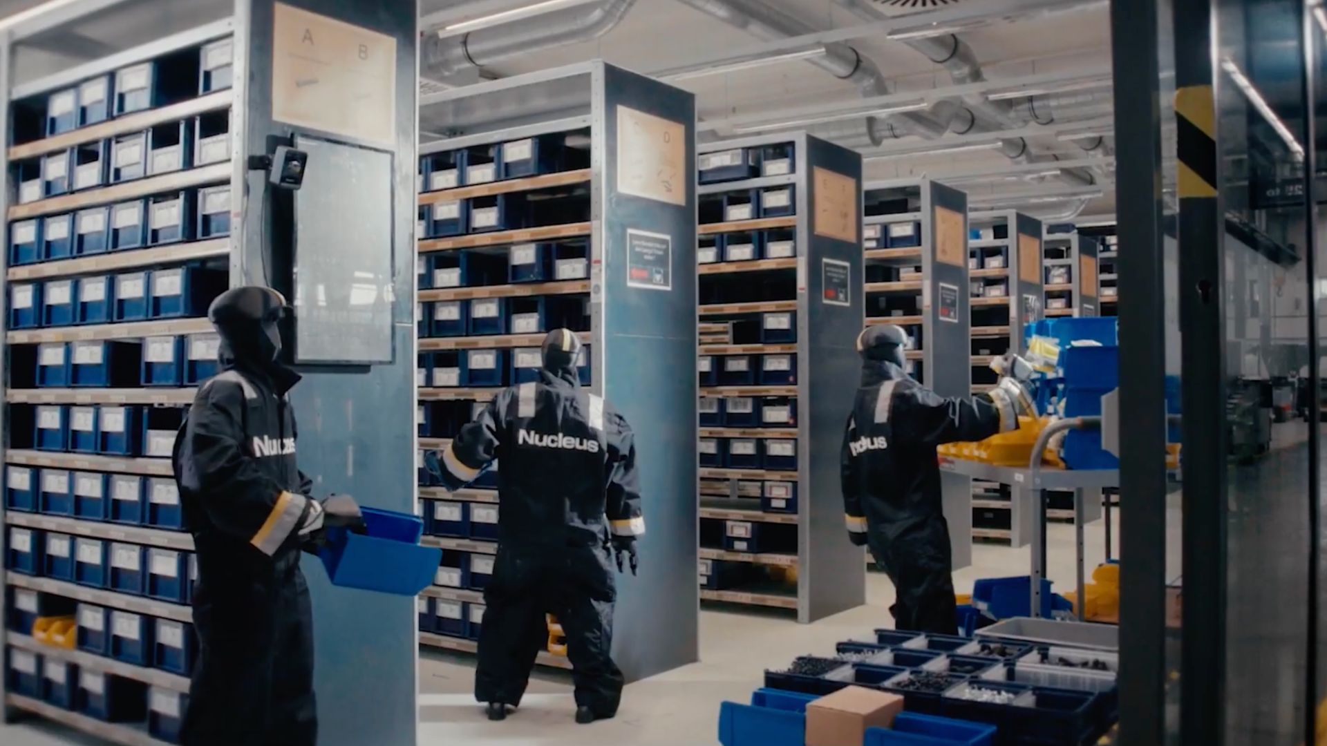 Three Nucleus-branded humanoid robots handling blue bins in a parts warehouse