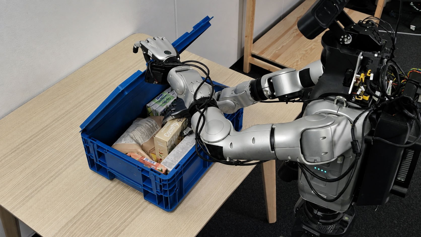 Silver dual robot arms packing groceries into a blue tote on a wooden table