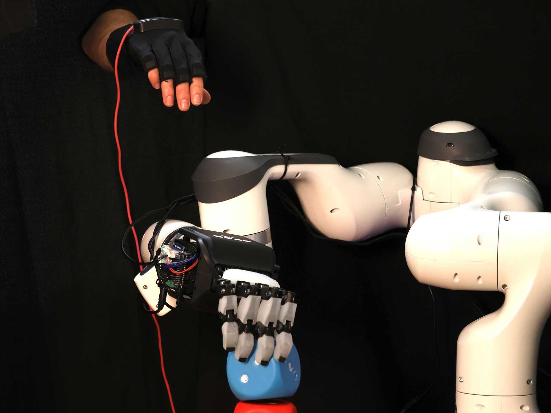 ORCA robotic hand on a robot arm grasping a blue cube while a person teleops with a glove