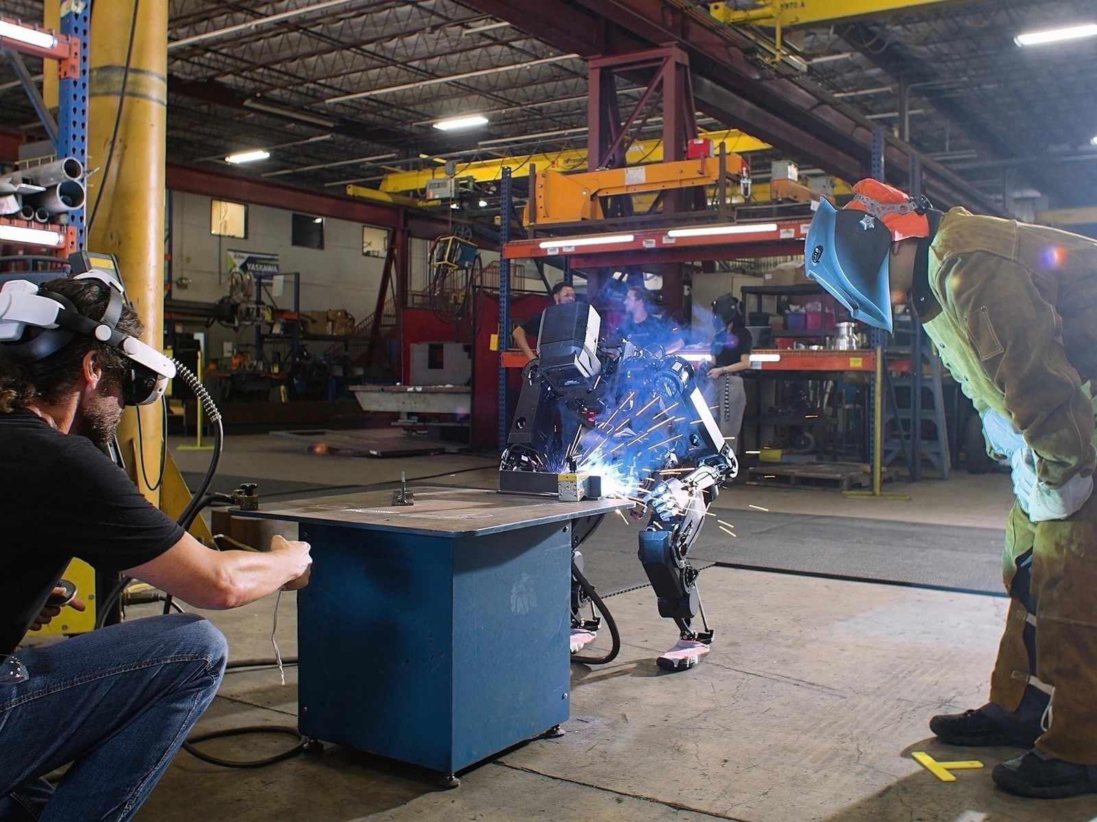 Persona AI humanoid welding at a steel table while an operator in a VR headset teleoperates and a welder in gear watches
