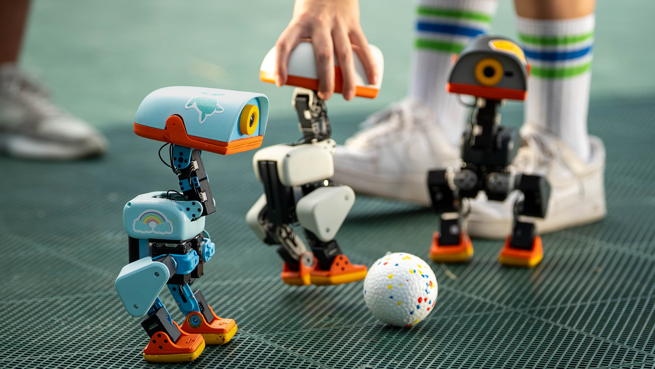 Three Microduck bipeds on a turf pitch around a small ball, one held by a hand