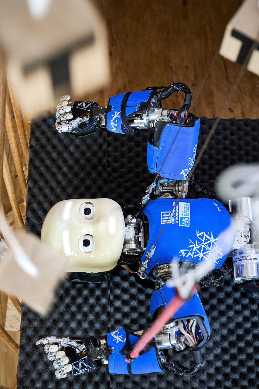 Close-up of iCub’s face and arms in the crib, a pink tether on one wrist, hanging blocks out of focus