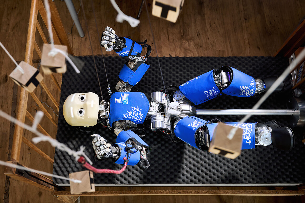 iCub humanoid lying on its back in a wooden crib, limbs tethered with strings to hanging blocks