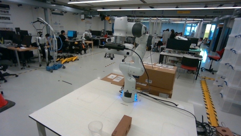 White Franka arm on a lab table with a cup and block, other robots in the background