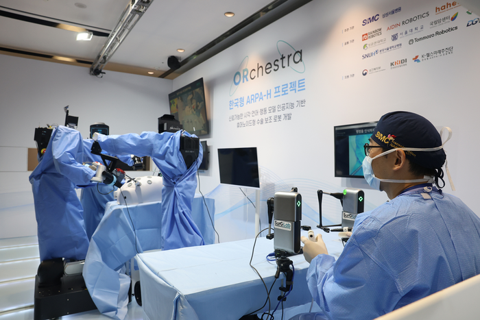 Surgeon at a draped table with two blue-gowned wheeled robot torsos and ORchestra partner logos on the wall