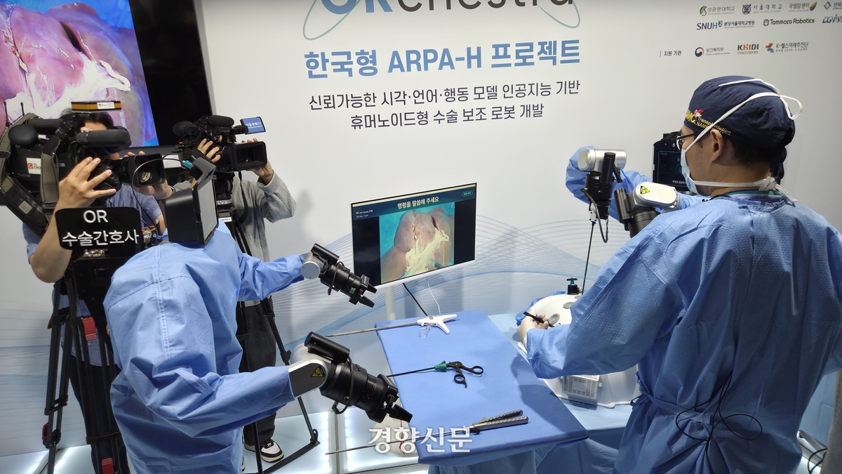 Rear view of two blue-gowned robots at a draped table with laparoscopic tools and a live endoscopic feed on a monitor