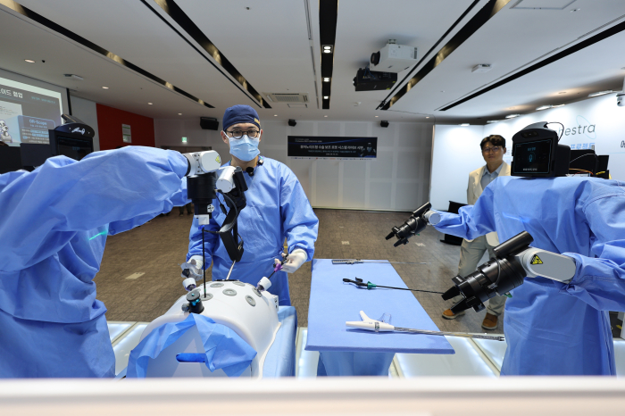 Two wheeled humanoid torsos in surgical gowns flanking a surgeon at a mock abdomen, with instruments laid out on a side table
