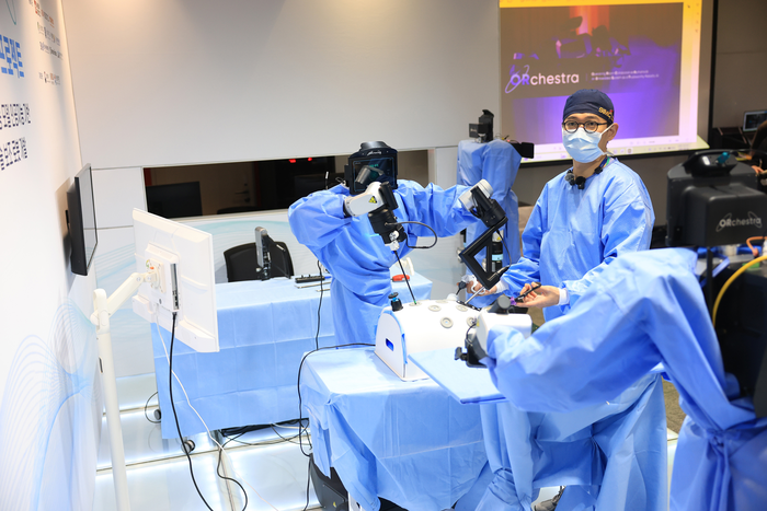 Surgeon in blue scrubs at a draped table with two blue-gowned humanoid arms assisting a mock laparoscopic case