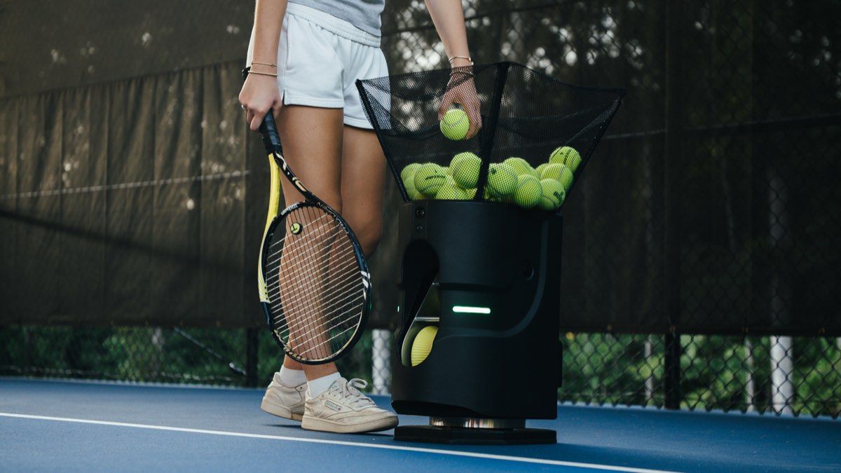 Person loading tennis balls into a Tennibot Partner Lite hopper on a blue court
