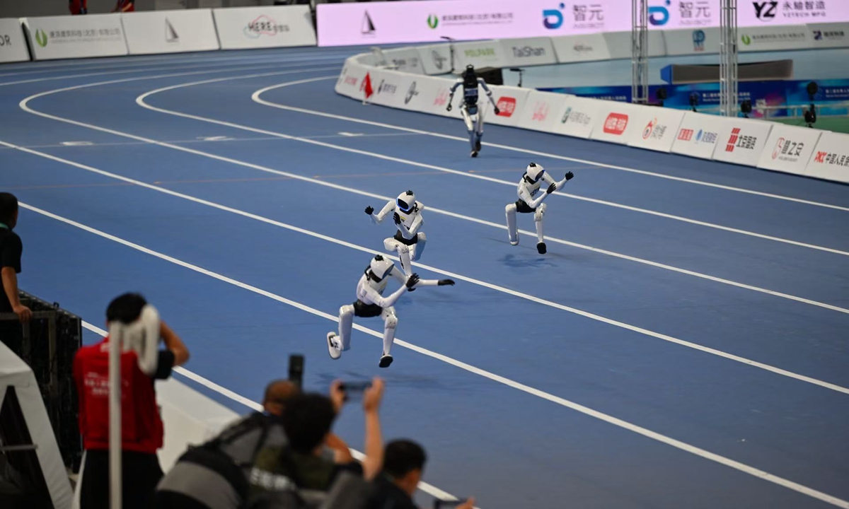 Four compact humanoids sprinting on a blue indoor track with arms held high near their heads