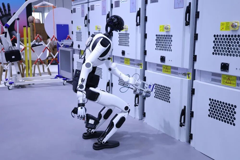 A compact white X-Humanoid standing at a wall of equipment cabinets, reaching a panel with one hand