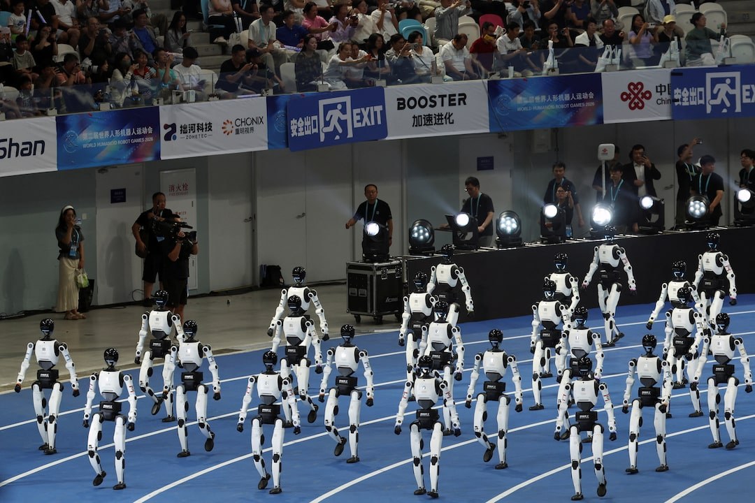 Rows of white Booster humanoid robots walking in formation on a blue indoor track