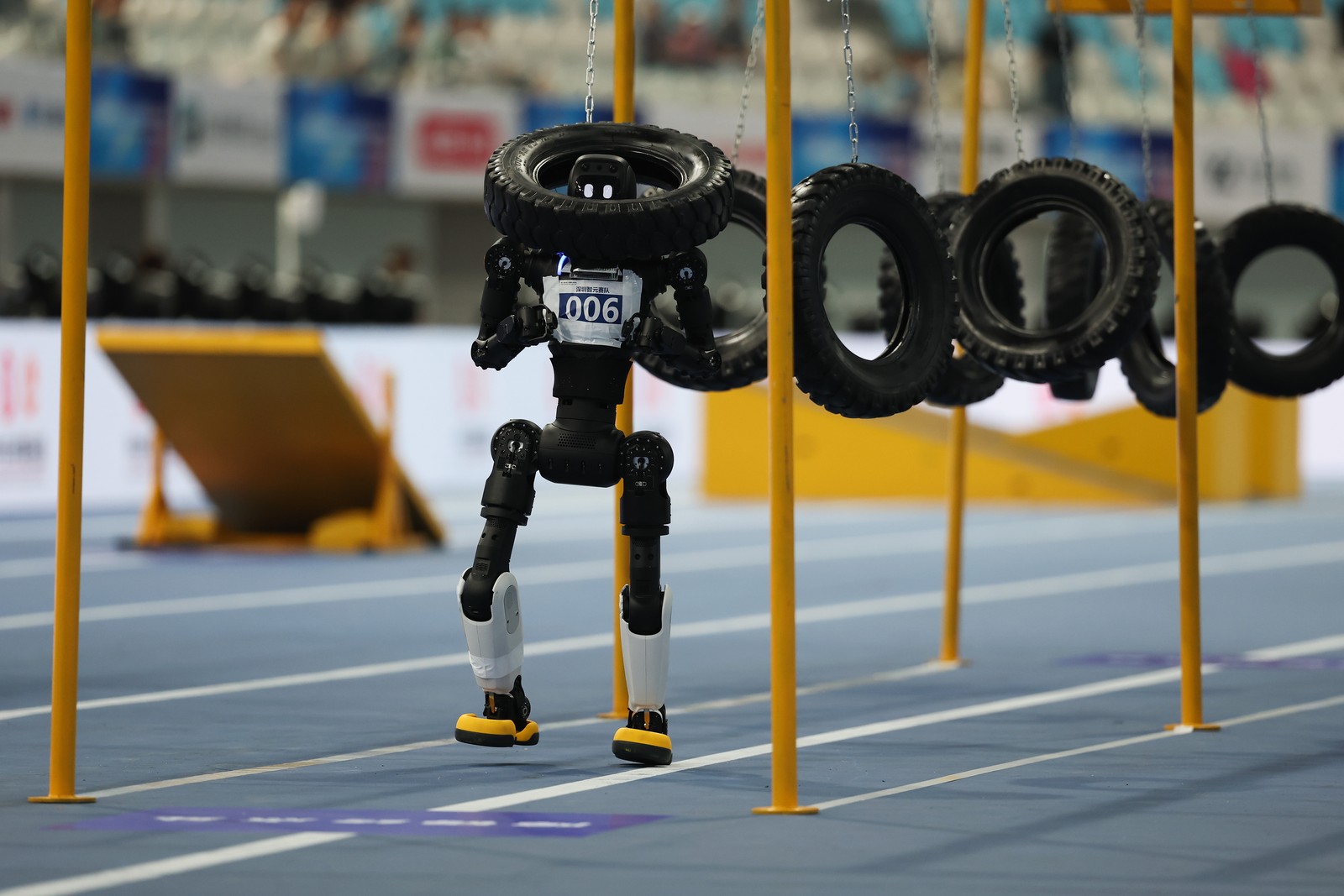A black humanoid robot with bib 006 walking through hanging tires on a blue indoor track