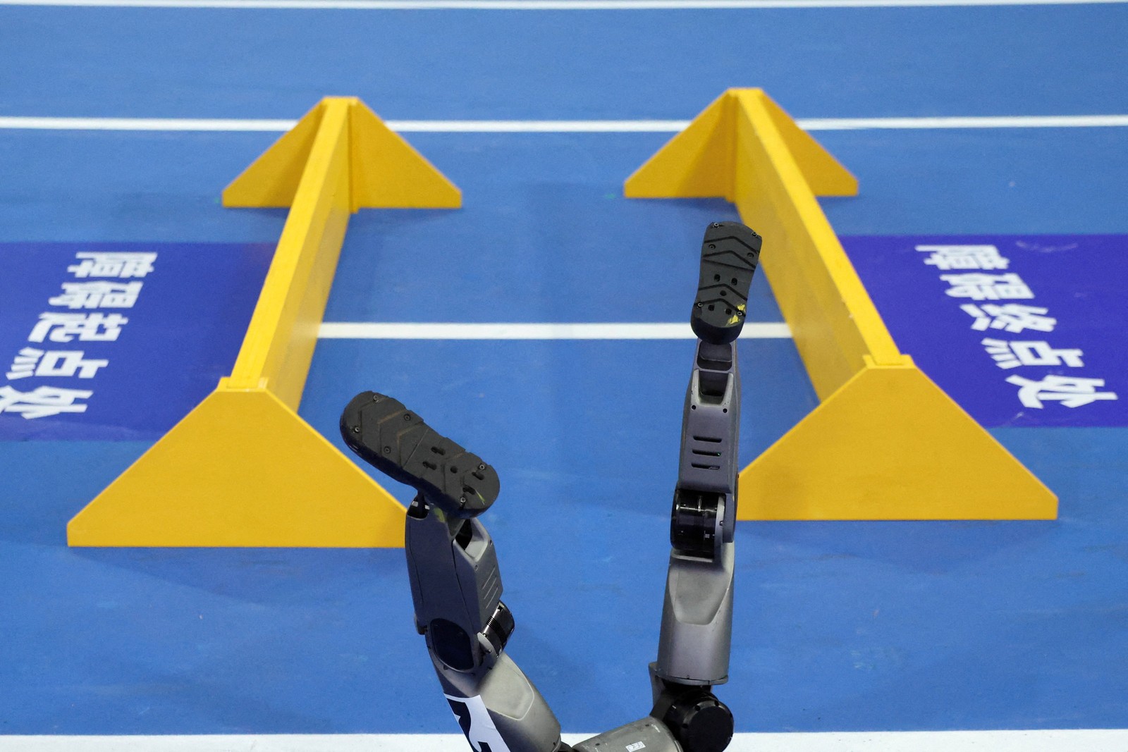 A fallen humanoid robot’s feet pointing up in front of yellow barriers on a blue track