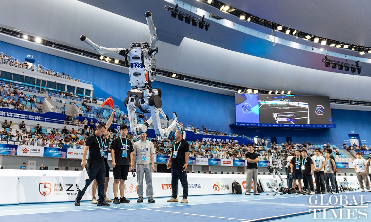 Humanoid robot bib 023 fully airborne over a blue indoor court with officials watching