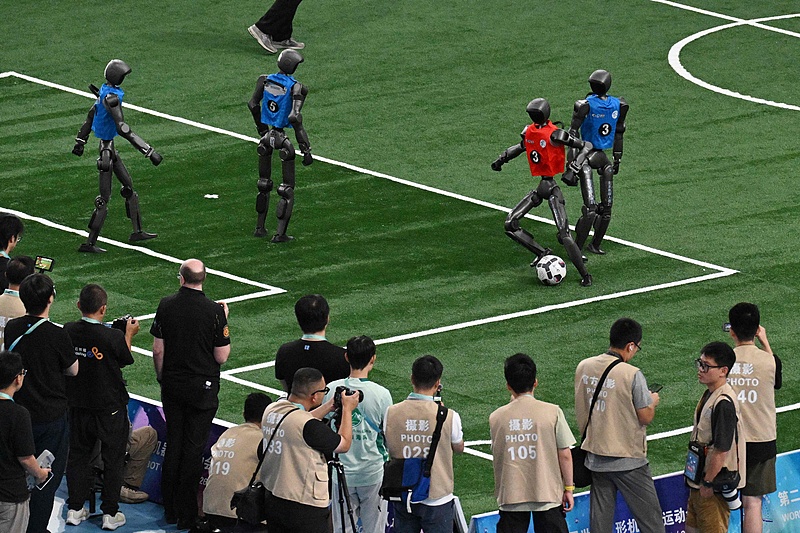 Full-size humanoid soccer robots on a turf pitch, a red-bib player dribbling past a blue-bib defender as photographers watch