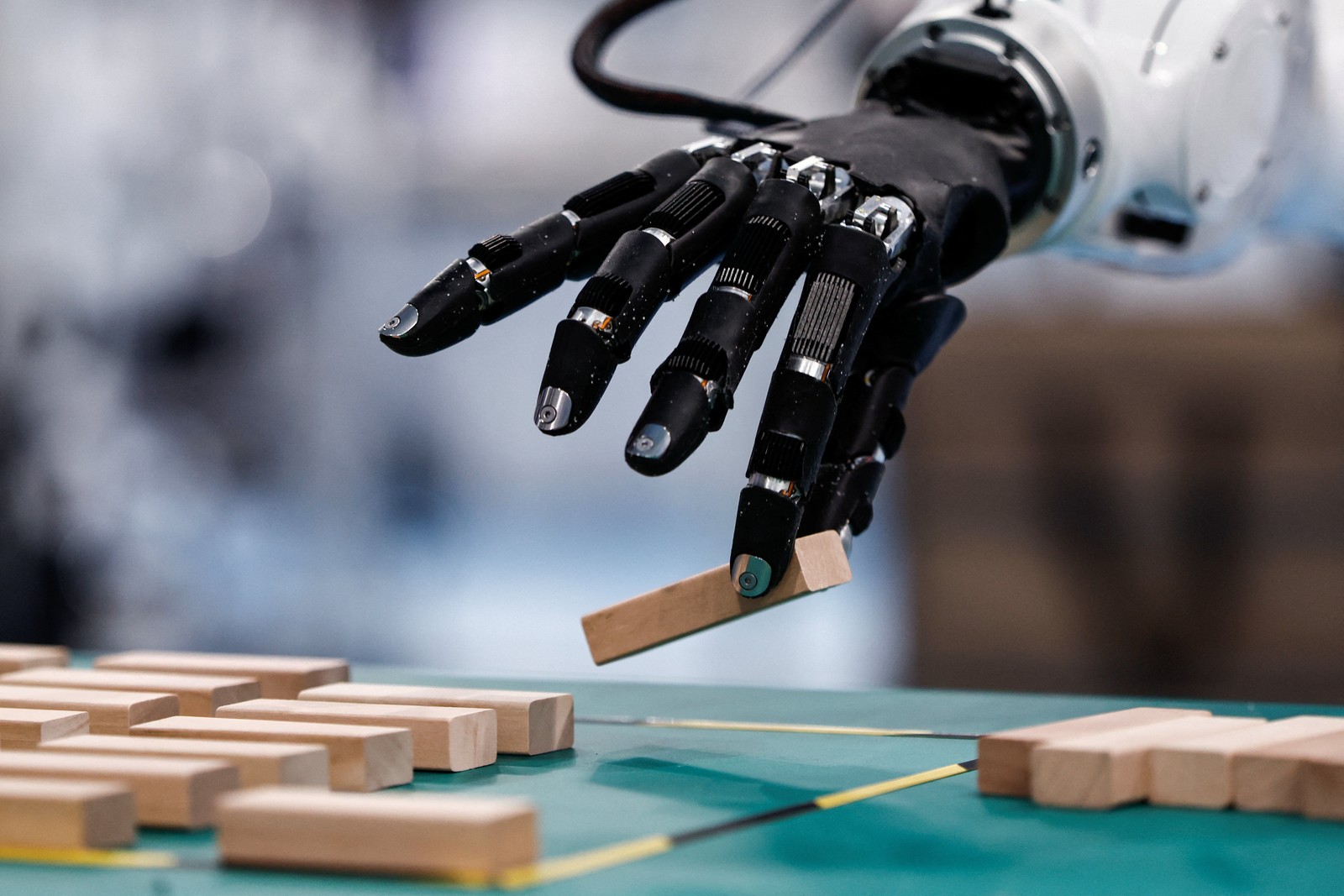 A black five-fingered robotic hand pinching a wooden block over a green table of identical blocks
