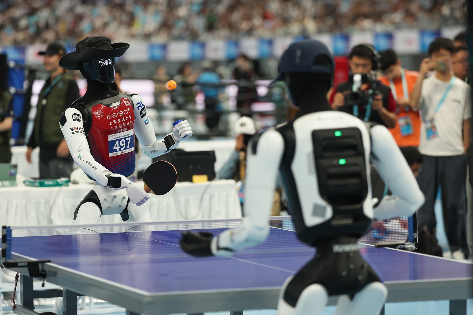 Two humanoid robots play table tennis; an orange ball hangs between them over a blue table