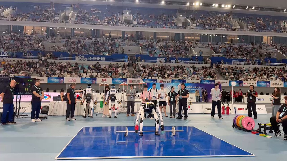 A humanoid bent over a barbell on the blue lifting platform with officials and other robots watching