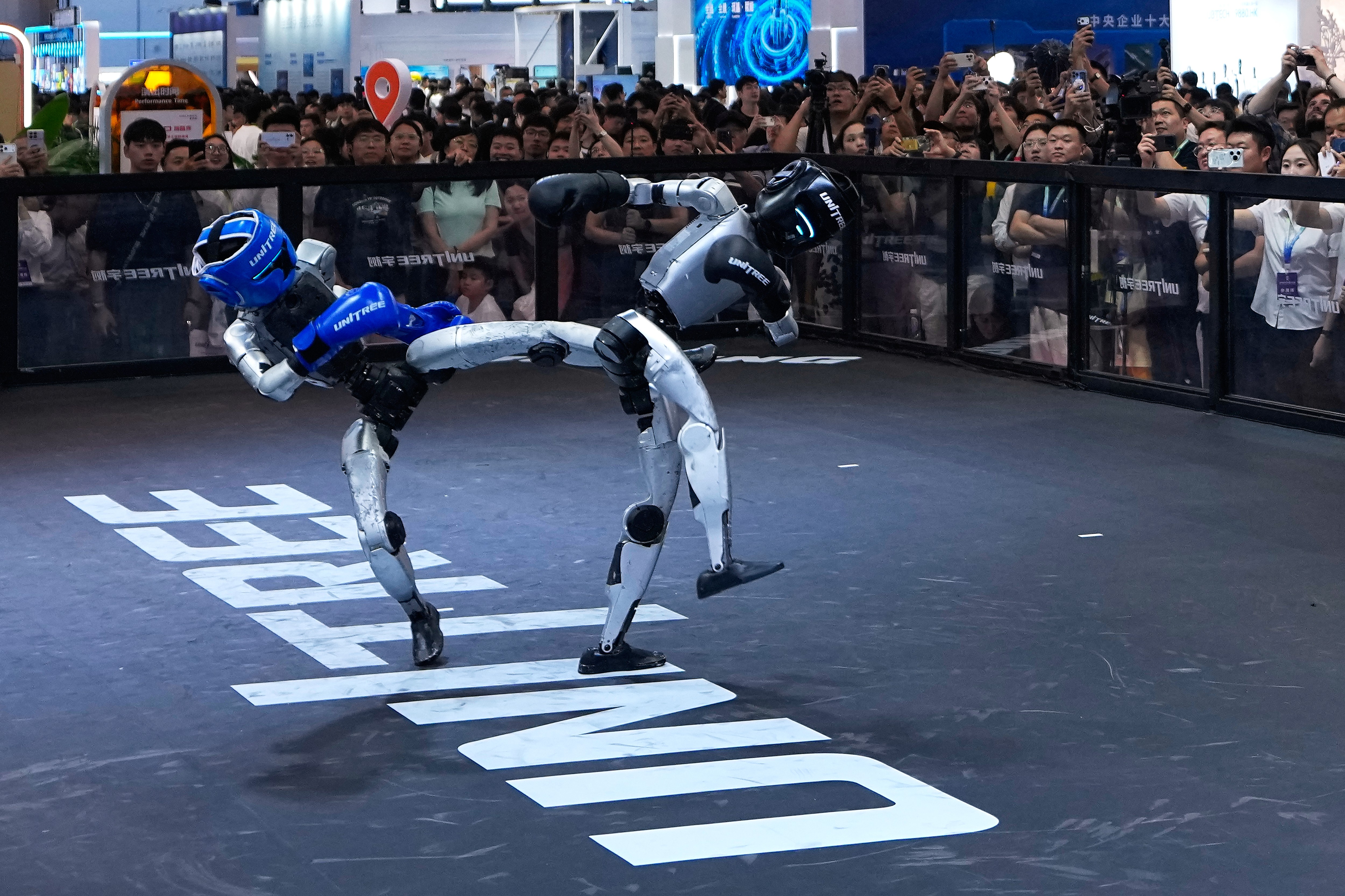 Two Unitree humanoid robots boxing in a ring as a crowd watches at the World Robot Conference in Beijing